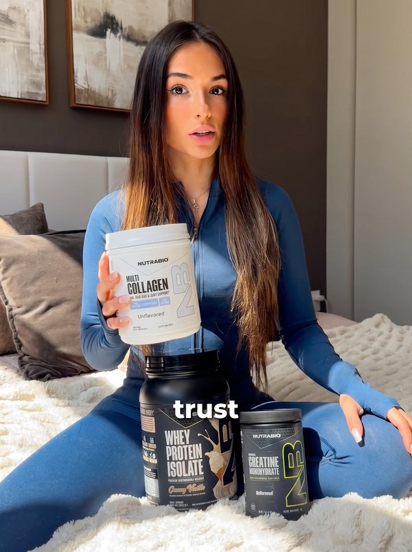 Creator on a bed holding Nutrabio collagen, with protein and creatine tubs in front of her